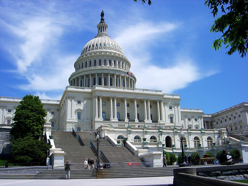The United States Capitol Building sits in the forefront against a blue sky with light, wispy clouds. The building frames the marble structures dome and steps.