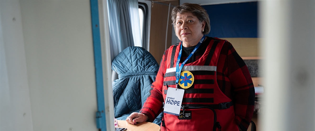 A photograph of a Project HOPE-supported health worker as she looks at the camera. She is on a mobile medical unit in Ukraine and wear a lanyard and bullet proof vest with a medical worker patch.